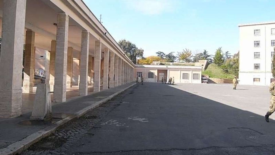 Ampio cortile asfaltato con un lungo porticato a sinistra, caratterizzato da una fila di colonne in muratura e soffitto piatto. Sullo sfondo si vedono edifici bassi e un palazzo a più piani, con alberi oltre il complesso sotto un cielo sereno.
