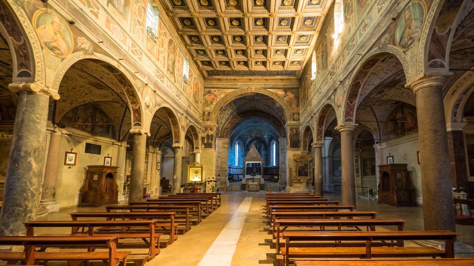 Interno di una chiesa storica con navata centrale fiancheggiata da colonne e archi, pareti affrescate e soffitto a cassettoni decorato. File di panche in legno conducono verso l’altare sul fondo, illuminato da finestre laterali.