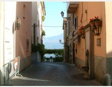 Vicolo stretto in ciottoli tra edifici dai colori chiari, con lampioni a parete e balconi con vasi di fiori. In fondo alla strada si intravede un lago con montagne sullo sfondo.