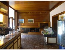 Interno di un piccolo bar con rivestimenti in legno su pareti e soffitto, bancone lungo sulla sinistra e pavimento in marmo scuro. Sul fondo c’è un grande camino incassato con quadri sopra, mentre a destra si vedono una vetrina espositiva e un frigorifero verticale per bevande.