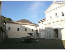 Piazza lastricata in pietra con una fontana circolare al centro, circondata da edifici chiari con finestre e tetti in tegole. Sullo sfondo si vede una collina boscosa sotto un cielo sereno.