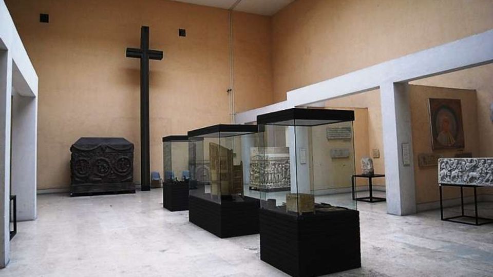 Sala museale con pareti beige e soffitto alto, illuminata da un lucernario, con tre teche in vetro su basi nere al centro. Sul fondo si vede una grande croce nera e un grande sarcofago scolpito, con altre opere esposte su piedistalli lungo le pareti.