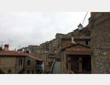 Veduta di un borgo collinare con case in pietra e tetti in coppi rossi, disposte su pendii e vicoli stretti. In primo piano si vede una strada in salita tra edifici, con lampione, antenne e camini sotto un cielo nuvoloso.