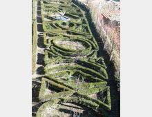 Giardino formale visto dall’alto con siepi di bosso potate a labirinto e aiuole interne, con una piccola fontana e vasca d’acqua nella parte superiore. Il giardino è affiancato da vialetti e confina con un edificio in pietra sul lato destro.