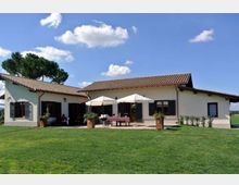 Casa rurale a un piano con tetto in coppi, persiane scure e veranda centrale con due ombrelloni e tavolo. Davanti si estende un ampio prato verde con vasi di piante e alberi sullo sfondo sotto un cielo azzurro con nuvole.