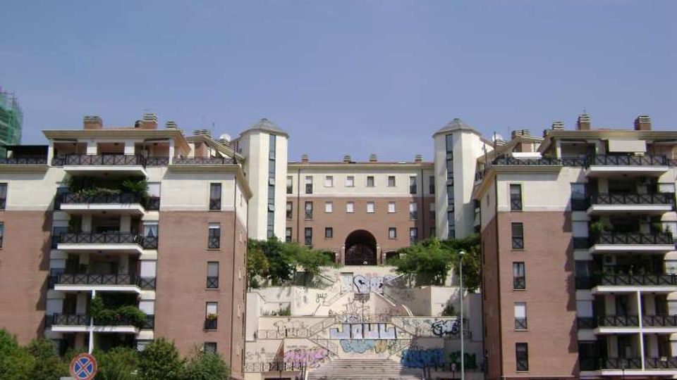 Complesso residenziale moderno con due palazzi in mattoni e cemento ai lati, dotati di balconi, e un edificio centrale più alto sullo sfondo. In primo piano una scalinata ampia con murales sui muri di contenimento conduce a un arco d’ingresso, con alberi e strada alla base.