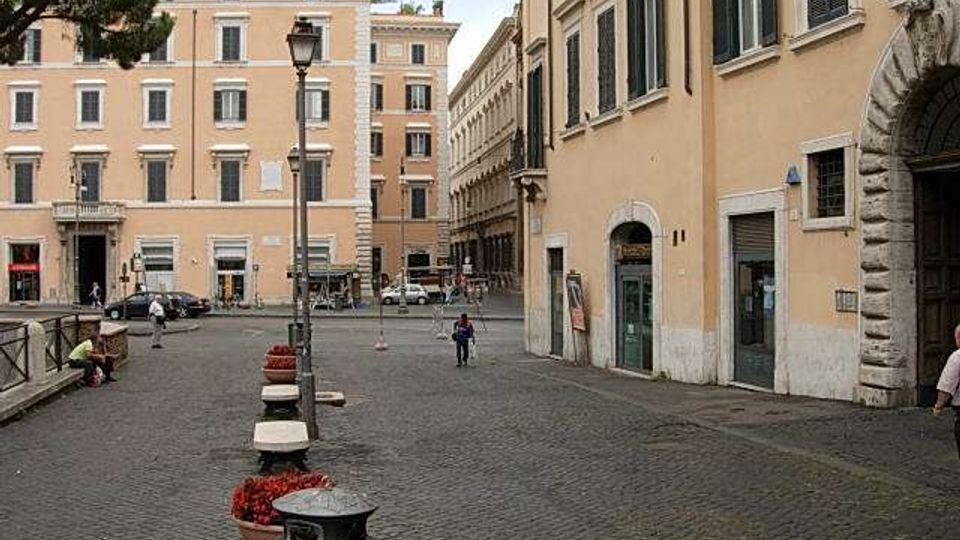 Piazza urbana con pavimentazione in sampietrini, lampioni e fioriere lungo un marciapiede, circondata da palazzi storici color ocra con finestre e persiane. Sullo sfondo si vedono facciate con cornici decorative e un’area stradale con auto parcheggiate.