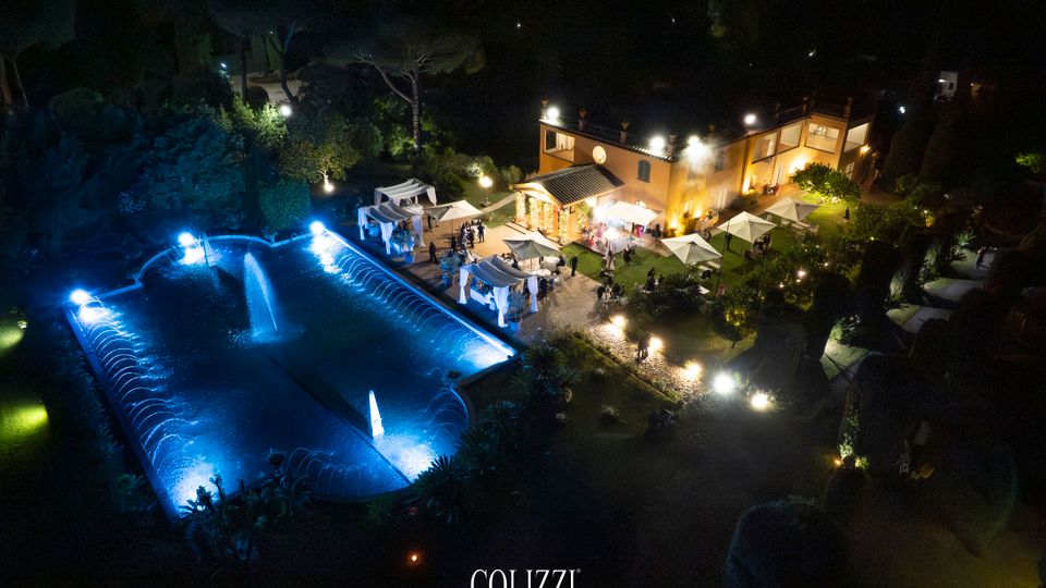 Veduta notturna dall’alto di una villa con giardino illuminato, area pavimentata e gazebo bianchi. In primo piano c’è una grande piscina rettangolare con fontane e luci blu, circondata da alberi e vialetti.