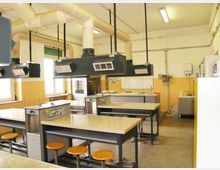 Aula laboratorio con grandi banchi da lavoro piastrellati e sgabelli, dotata di cappe aspiranti sospese e tubazioni di ventilazione a soffitto. Lungo le pareti sono presenti finestre, lavelli e attrezzature tecniche tipiche di un laboratorio scolastico o scientifico.