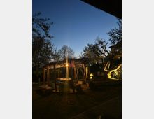 Giardino al crepuscolo con pergolato in legno e area tavoli illuminata da luci calde. Alberi e vegetazione circondano lo spazio, con decorazioni appese e cielo blu scuro sullo sfondo.