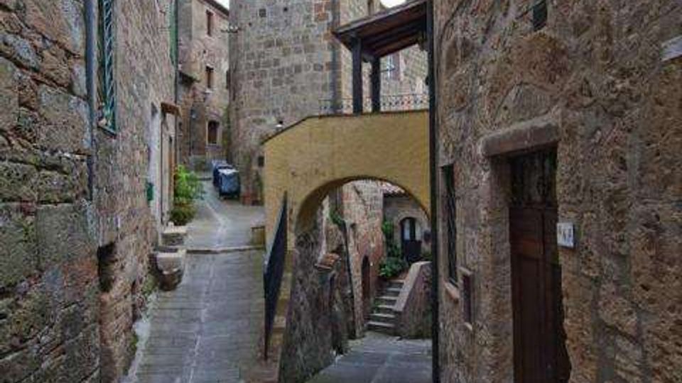 Vicolo stretto in un borgo medievale con edifici in pietra e pavimentazione lastricata, che si biforca tra una rampa e una scalinata. Un arco-ponte intonacato collega due costruzioni sopra il passaggio, con una torre in pietra sullo sfondo.