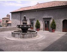 Cortile in pietra con fontana ottagonale in stile antico al centro, su pavimentazione lastricata. Sullo sfondo un edificio in muratura di pietra con tetto in coppi e porte in legno, affiancate da due vasi con arbusti.