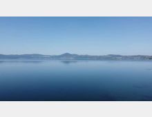 Vista panoramica di un grande lago con acqua calma e riflettente sotto un cielo sereno. All’orizzonte si vedono colline e una sponda lontana con edifici sparsi.