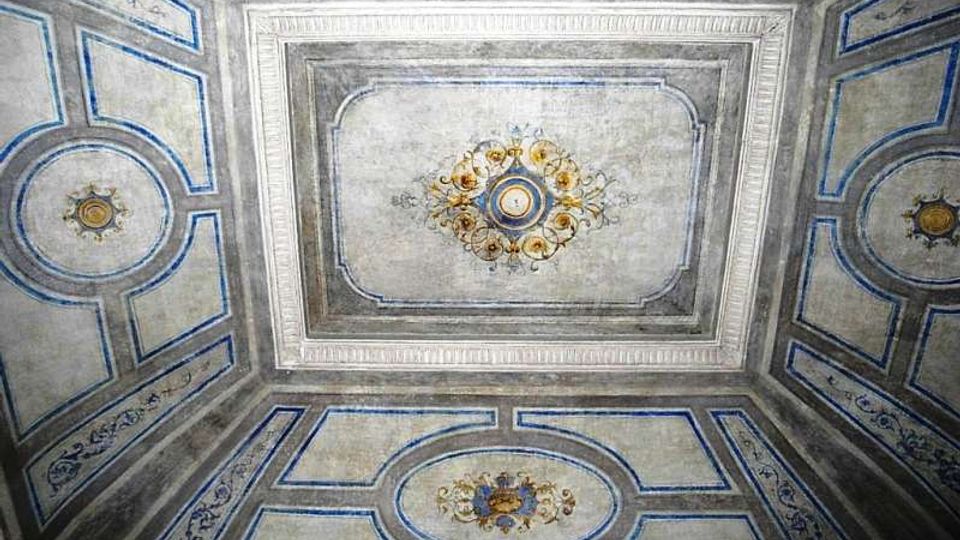 Soffitto interno decorato con riquadri geometrici bordati in blu e cornici in stucco, con pannello centrale rettangolare e medaglioni ornamentali dorati. La superficie è dipinta in tonalità chiare con motivi floreali e rosoni simmetrici.