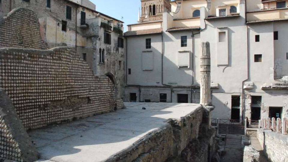 Resti archeologici in pietra con una lunga passerella sopraelevata e un alto muro a mosaico sul lato sinistro. Sullo sfondo si vedono edifici storici con facciate intonacate e un campanile in mattoni che svetta sopra i tetti.