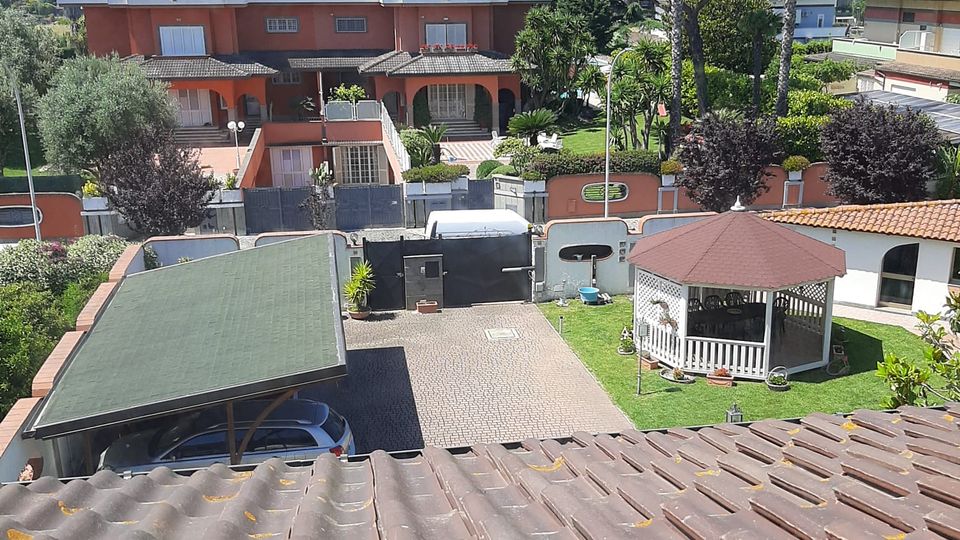 Vista dall’alto su un quartiere residenziale con villette dai tetti in tegole, un vialetto pavimentato con carport e un giardino con gazebo bianco. Sullo sfondo si vedono edifici a due piani, alberi e alte palme sotto un cielo sereno.