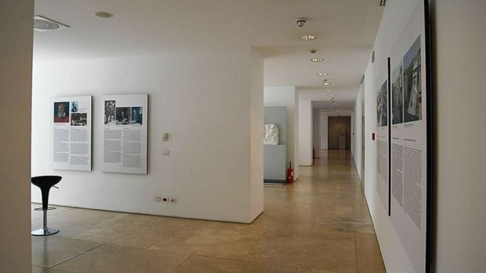 Interno di una galleria o museo moderno con pareti bianche e pavimento in grandi lastre di pietra, illuminato da faretti incassati nel soffitto. Lungo le pareti sono esposti pannelli informativi e fotografie, con un corridoio che si estende in profondità verso altre sale.