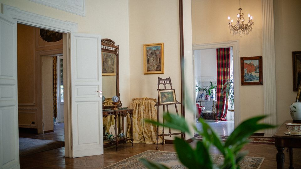 Interno di una casa in stile classico con pavimento in parquet e porte bianche alte che collegano più stanze. Sono visibili un lampadario a bracci, quadri alle pareti, un piccolo tavolo con specchiera, tappeti e una finestra con tende a righe rosse e verdi accanto a piante in vaso.