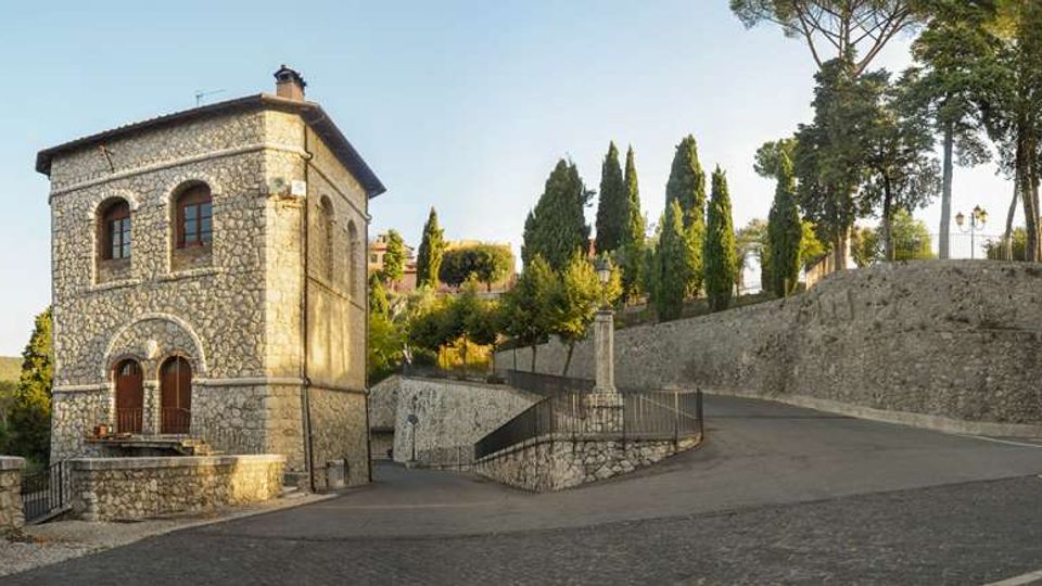 Edificio in pietra a due piani con archi e finestre ad arco, affacciato su una strada in salita. Accanto si vedono mura in pietra, una scalinata con ringhiera e alberi alti (cipressi e pini) lungo il pendio.
