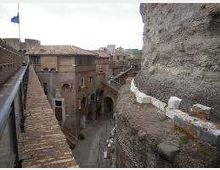 Vista da una passerella in pietra lungo una parete rocciosa, con parapetto e bandiera, che conduce verso un complesso di edifici storici in muratura e un arco su una strada sottostante. Sullo sfondo si vedono tetti in coppi e mura in pietra in un contesto di borgo antico.