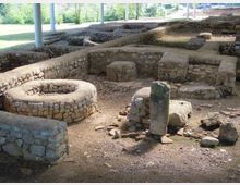 Scavi archeologici all’aperto con muri bassi in pietra che delineano ambienti rettangolari e un’area circolare, sotto una tettoia sostenuta da pilastri. Il terreno è in terra battuta con pietre sparse, e sullo sfondo si vede un’area verde con alberi.