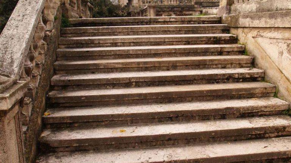 Scalinata esterna in pietra con balaustre scolpite che conduce a un edificio monumentale in stile classico con facciata simmetrica e finestre rettangolari. Lungo la scalinata sono presenti lampioni e vegetazione ai lati.