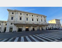 Facciata di una stazione ferroviaria storica a tre piani, con intonaco chiaro, finestre alte e un lungo portico con colonne e archi d’ingresso. Davanti si vede una strada con attraversamento pedonale a strisce e cavi aerei sopra la carreggiata.