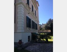 Facciata laterale di una villa storica intonacata color crema, con persiane scure e cornici in rilievo alle finestre. In primo piano si vede un cortile pavimentato in pietra con vaso ornamentale e, sullo sfondo, una balaustra e un giardino con alberi.