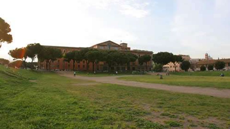 Ampio prato urbano con sentieri sterrati e filari di pini marittimi, con un lungo edificio storico in mattoni sullo sfondo. Cielo aperto e spazi verdi estesi che definiscono un’area di parco o piazza.