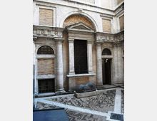 Cortile interno di un edificio storico con facciata in pietra e mattoni, colonne e archi, e una grande finestra con grata in ferro. Sul pavimento in pietra decorato è visibile una scultura sdraiata su un basamento, vicino a un ingresso laterale.