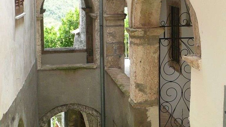 Vicolo stretto tra edifici in muratura chiara con porticati ad archi in pietra e un cancelletto in ferro battuto, con piccoli gradini e piante in vaso. In fondo si vedono altre arcate e uno scorcio di vegetazione e colline sullo sfondo.