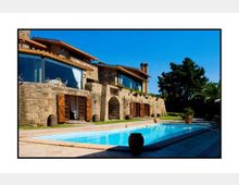 Villa in pietra a più livelli con grandi vetrate e porte in legno, affacciata su una lunga piscina rettangolare. Ampio patio pavimentato con vasi e giardino con alberi sullo sfondo sotto cielo sereno.