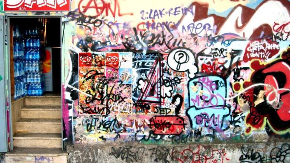 Ingresso di un bar con insegna “BAR” e gradini che scendono verso una porta aperta, con scaffali di bottiglie visibili all’interno. Accanto, un grande muro esterno ricoperto di graffiti multicolori, scritte e poster pubblicitari sovrapposti.