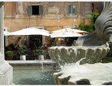 Fontana in pietra in una piazza, con vasca d’acqua in primo piano e schizzi visibili. Sullo sfondo, un edificio storico con finestre e tende da esterno bianche sopra tavoli di un’area caffè.