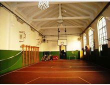 Palestra scolastica al coperto con campo segnato a terra e canestro da basket su una parete di fondo. Soffitto alto con travi a vista, pareti bianche e verdi, spalliere in legno e grandi finestre laterali.