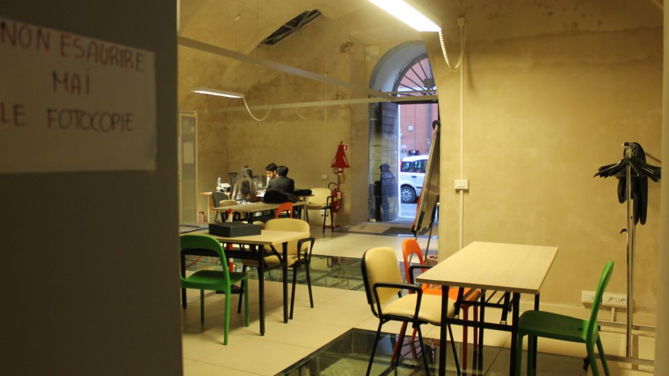 Interno di una sala studio in un edificio con pareti in intonaco grezzo e soffitto a volta, illuminata da lampade lineari. Sono presenti tavoli con sedie di colori diversi, un pavimento in piastrelle con inserti in vetro e un grande portale ad arco che si apre verso l’esterno.