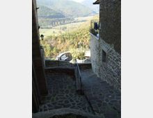 Scalinata e terrazzamento in pietra tra edifici in muratura, con ringhiere metalliche e lampione, affacciati su una valle verde con boschi e campi ondulati. Sullo sfondo si vedono colline boscose che chiudono il panorama.