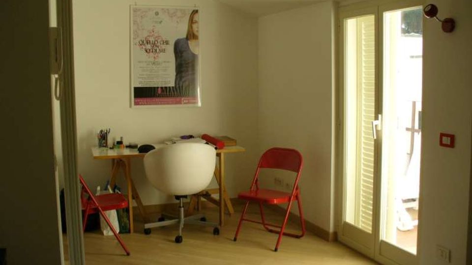 Piccolo studio con pavimento in legno chiaro, tavolo da lavoro con sedia bianca e due sedie pieghevoli rosse, e un poster appeso alla parete. Una porta-finestra con persiane lascia entrare molta luce e dà accesso a un balcone.