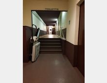 Corridoio interno con una breve rampa di scale e corrimano, illuminato da luci a parete e con soffitto in legno scuro. In alto è visibile la scritta “TOILETTE” sopra l’accesso, con porte lungo il lato destro e un dislivello verso l’area dei bagni in fondo.
