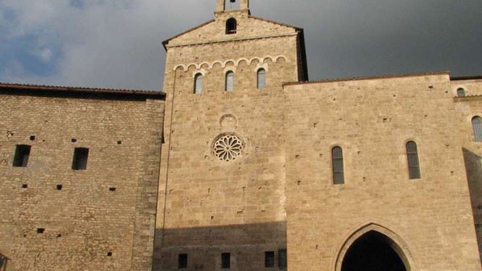 Facciata in pietra di una chiesa medievale con campanile a vela, rosone centrale e serie di archetti sotto la sommità. In basso si vede un grande portale ad arco ogivale e finestre strette, con un’ampia parete in pietra sul lato.