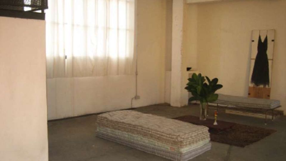 Interno di un ampio loft con pareti bianche e pavimento in cemento, illuminato da una grande finestra con tende leggere. Nella stanza ci sono un materasso basso a terra, una panca metallica, un tappeto con una pianta in vaso e un lampadario al soffitto.