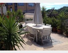 Terrazza pavimentata all’aperto con tavolo ovale e sedie bianche in plastica, ombrellone chiuso al centro e lettini sullo sfondo. Numerose piante in vaso con foglie appuntite circondano l’area, con edifici e tetti visibili oltre la ringhiera.