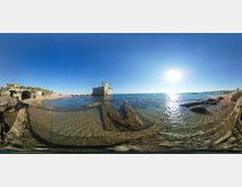 Panorama di una costa rocciosa con acqua bassa e trasparente; al centro si vede una piccola torre in pietra su uno scoglio vicino alla riva. Sulla sinistra ci sono scogli e vegetazione costiera, mentre a destra si estende il mare aperto con il sole basso che si riflette sull’acqua.