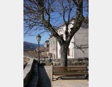 Terrazza panoramica in un borgo collinare con panchina, grande albero spoglio e lampione in ferro accanto a un muretto in pietra. Sullo sfondo si vedono case in muratura con persiane e tetti in coppi, con montagne all’orizzonte sotto cielo sereno.