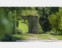 Sentiero in un parco verde con prato e alberi, con un vecchio arco/portale in pietra coperto da rampicanti al centro della scena. Vegetazione fitta e cespugli incorniciano il percorso in una giornata luminosa.