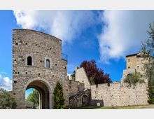 Porta fortificata in pietra con torre semicircolare e grande arco d’ingresso, collegata a mura merlate con una scala laterale. Sullo sfondo si vedono alberi e un edificio in pietra sotto un cielo azzurro con nuvole.