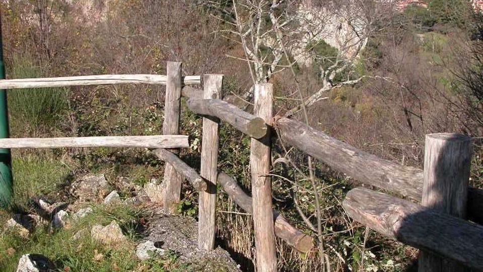 Vista panoramica di un borgo medievale arroccato su una collina rocciosa, con case in pietra addossate tra loro e una torre in cima. In primo piano si vede un sentiero con staccionata in legno e vegetazione, con colline boscose sullo sfondo sotto un cielo sereno.