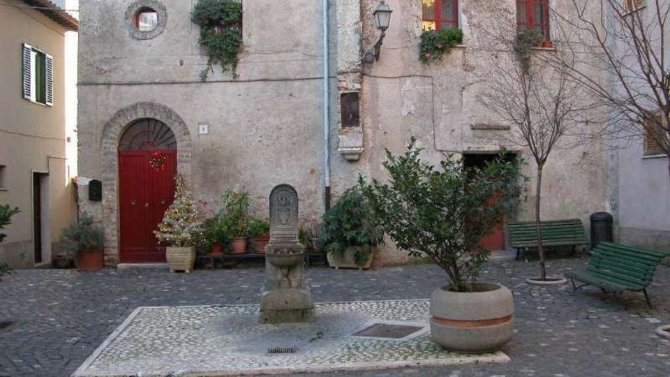 Piccola piazza lastricata in ciottoli con una fontanella in pietra al centro, piante in vaso e panchine verdi ai lati. Sullo sfondo, facciate di edifici in intonaco grezzo con porta ad arco rossa, finestre con persiane e una lanterna a parete.