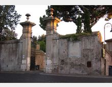 Ingresso con cancellata in ferro tra due alti pilastri in pietra sormontati da vasi ornamentali, inserito in un lungo muro intonacato e invecchiato. Sullo sfondo si vedono alberi alti e vegetazione sopra il muro, con strada asfaltata in primo piano.