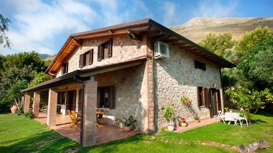 Casa di campagna in pietra a due piani con tetto a falde e persiane scure, dotata di portico coperto con colonne e patio. Intorno c’è un ampio prato con alberi, vasi e un tavolo da esterno, sullo sfondo colline o montagne sotto un cielo parzialmente nuvoloso.